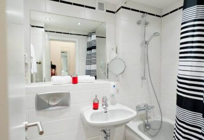 Bathroom at Hotel Esplanade Köln, a gay-friendly hotel. Features white tiles, sink, tub with shower, mirror, and black and white striped shower curtain.