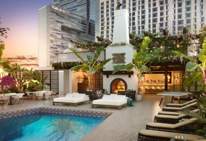 Hotel Figueroa, In The Unbound Collection By Hyatt gay-friendly hotel pool. Lounge chairs, fireplace, and bar area. Tropical plants adorn the space.