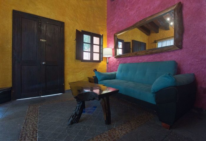 Hotel Hacienda Maria Bonita's gay-friendly room. A blue sofa sits beneath a rustic mirror. Yellow and pink walls add vibrant color.