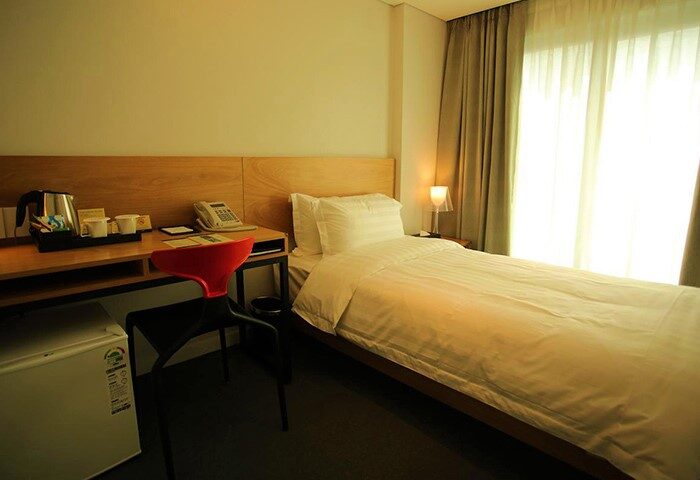 Hotel ITW gay-friendly room. Single bed with white linens, desk with kettle and phone, red chair, mini fridge. Window with curtains.