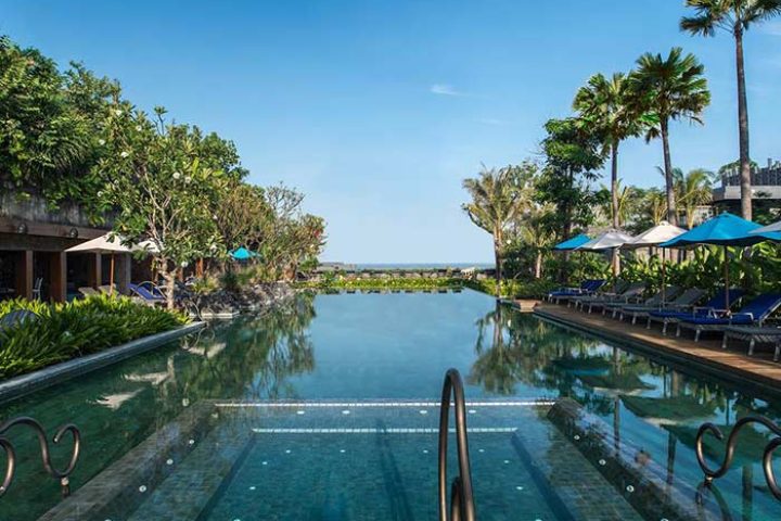 Hotel Indigo Bali Seminyak Beach pool view. Gay-friendly hotel with palm trees, lounge chairs, and ocean view. Ideal for gay travel.