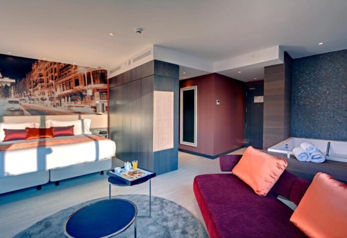 Hotel Indigo Madrid Gran Via room. Gay-friendly hotel with bed, couch, and jacuzzi. Cityscape mural behind the bed. Modern design.