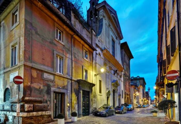 Street view of Hotel Indigo Rome - St. George, a gay-friendly hotel in Rome. Cobblestone street, historic buildings, and warm lighting.