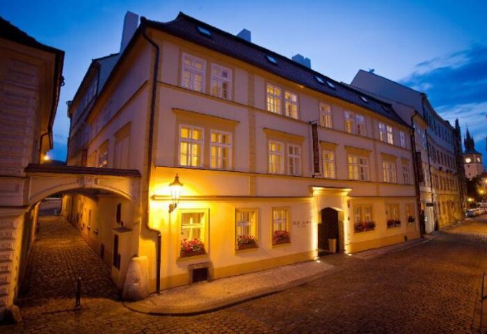 Hotel Leonardo Prague exterior at dusk. Gay-friendly, pet-friendly hotel with illuminated windows and cobblestone street. Prague travel.