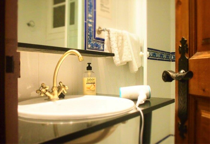 Bathroom at Hotel Liberty Sitges, a gay-friendly hotel. Sink, gold faucet, hand soap, and hair dryer on the counter. Part of a door visible.