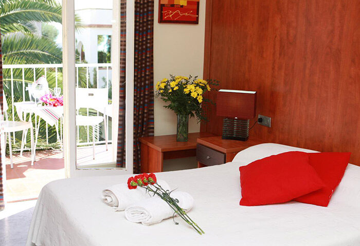 Hotel Los Globos gay-friendly room with balcony. Red pillows, towels, and flowers on the bed. Sunny, bright, and welcoming space.
