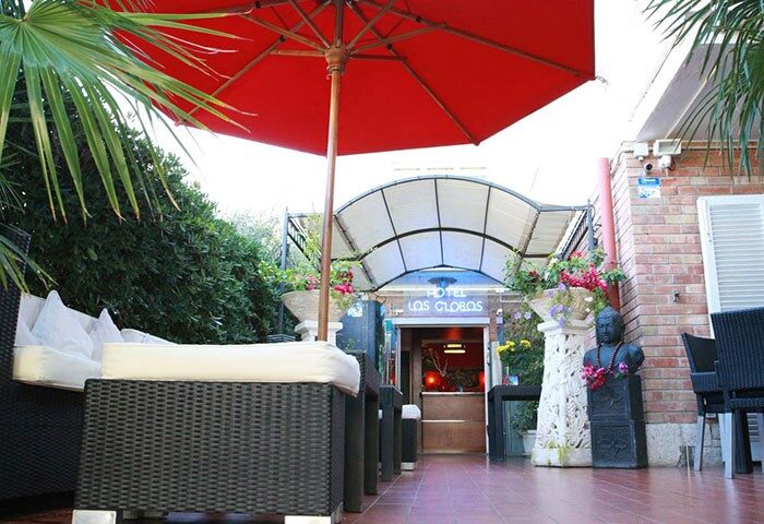 Hotel Los Globos, a gay-friendly hotel. View of the entrance with outdoor seating, red umbrella, and lush greenery. Gay travel destination.