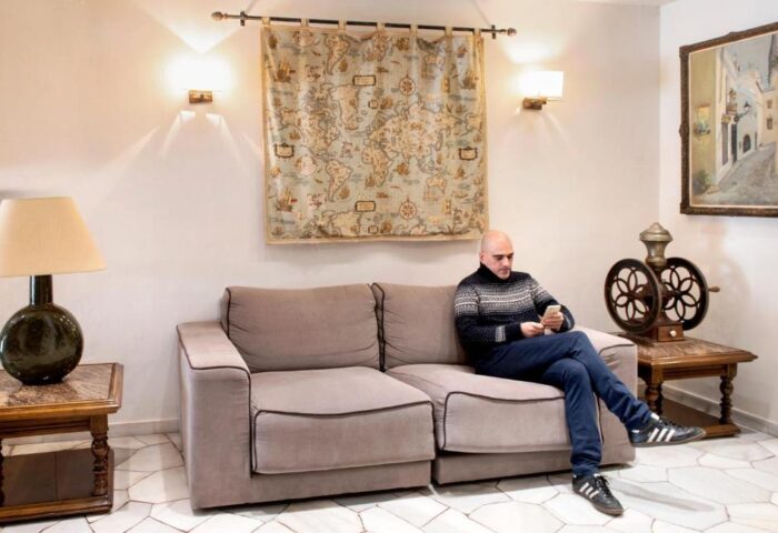 Hotel Los Globos gay-friendly space: man on couch using phone. Cozy interior with map tapestry, lamp, and vintage decor. Welcoming travel spot.