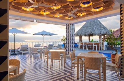Hotel Luxury Patio Azul's gay-friendly outdoor dining area. Tables, lounge chairs, umbrellas, and a thatched-roof bar overlook the ocean.