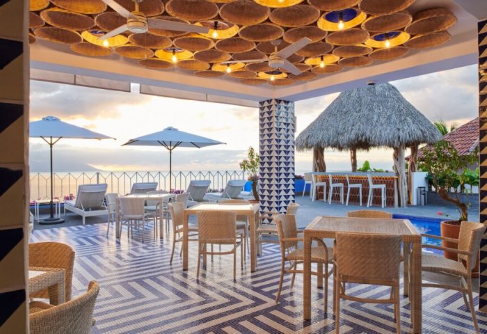 Hotel Luxury Patio Azul's gay-friendly outdoor dining area. Tables, lounge chairs, umbrellas, and a thatched-roof bar overlook the ocean.