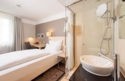 Hotel Lyskirchen gay-friendly room. Modern design with bed, desk, and bathroom featuring a glass-enclosed shower and sleek sink.