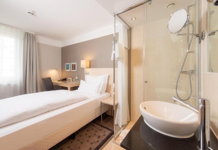 Hotel Lyskirchen gay-friendly room. Modern design with bed, desk, and bathroom featuring a glass-enclosed shower and sleek sink.