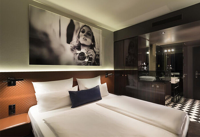 Hotel MANI by AMANO room. Gay-friendly hotel featuring a bed with white linens, a black & white photograph, and a bathroom with checkered floors.