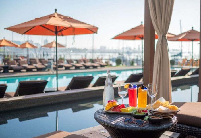 Poolside view at Hotel Maya A Doubletree Hotel, a gay-friendly hotel. Cocktails, fruit, and chips on a table by the pool. Umbrellas and lounge chairs visible.