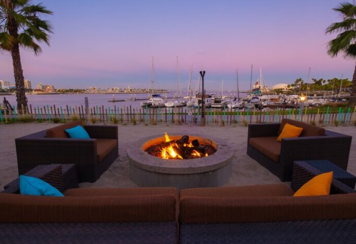 Hotel Maya A Doubletree Hotel's gay-friendly fire pit lounge area at dusk. Waterfront view, boats, palm trees, and cozy seating.
