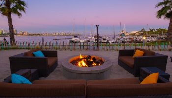 Hotel Maya A Doubletree Hotel's gay-friendly fire pit lounge area at dusk. Waterfront view, boats, palm trees, and cozy seating.