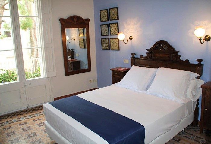 Hotel Medium Renaixenca gay-friendly room. Double bed with blue runner, wooden headboard, bedside tables, and lamps. Mirror and art on blue wall.