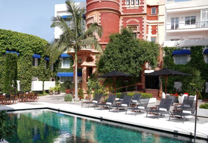 Pool view at Gay-Friendly Hotel Medium Sitges Park. Loungers line the pool's edge. Lush greenery and unique architecture surround the area.