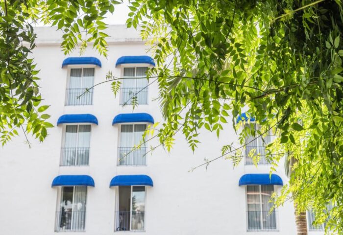 Facade of Hotel Medium Sitges Park, a gay-friendly hotel in Sitges. White building with blue awnings & green foliage. Gay travel advice.