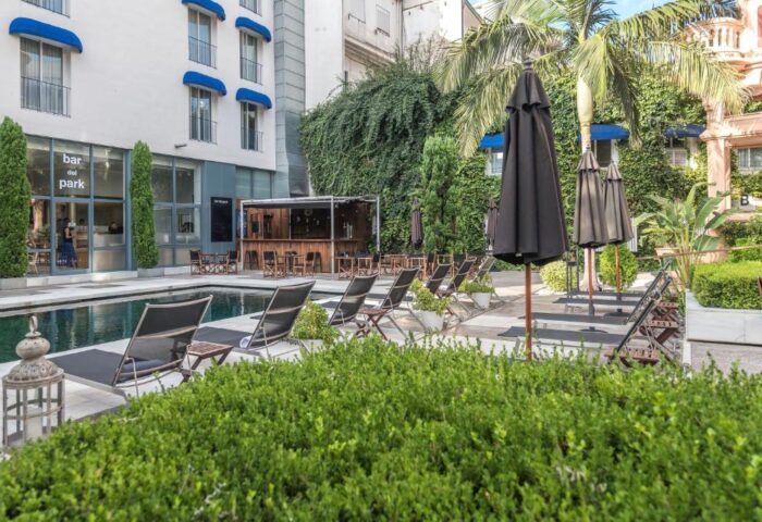 Hotel Medium Sitges Park pool area. Gay-friendly hotel with bar, lounge chairs, and umbrellas. Lush greenery surrounds the pool.
