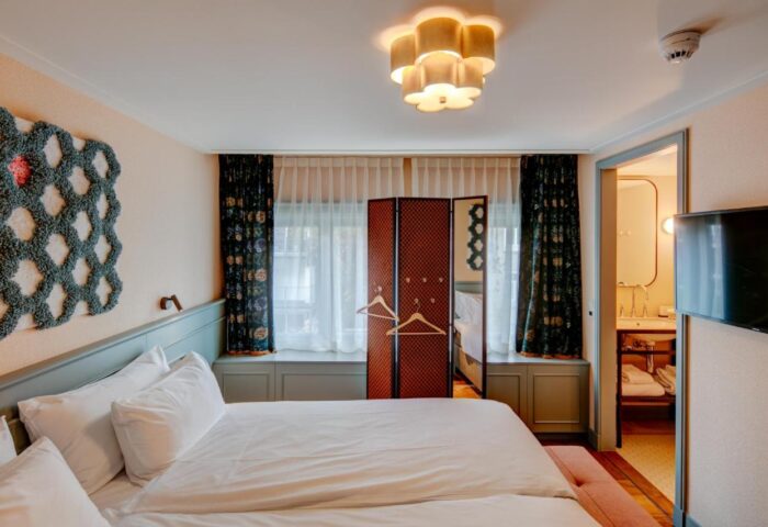 Hotel Mercier's gay-friendly room. A queen bed faces a decorative screen & bathroom. Floral curtains add style. A modern light fixture illuminates.