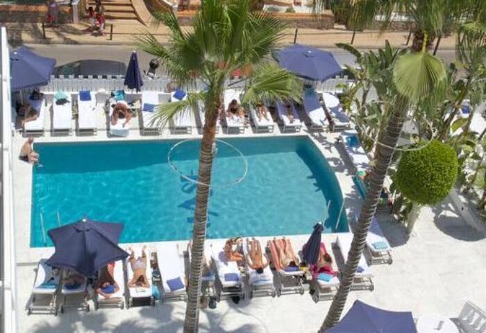 Hotel MiM Ibiza Es Vive Adults Only pool view. Gay-friendly hotel with sun loungers, blue parasols, and palm trees. Ideal for gay travel.