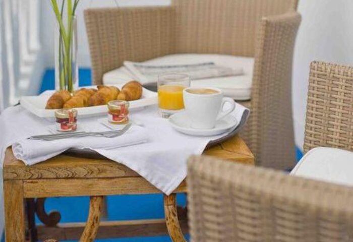 Breakfast at Hotel MiM Ibiza Es Vive, an adults-only, gay-friendly hotel. Croissants, juice, coffee, & jam on a table with wicker chairs.