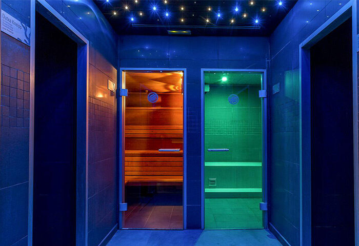 Hotel MiM Sitges gay-friendly hotel sauna. Two saunas with glass doors, one orange, one green, in a blue-lit room. Sauna benches visible.