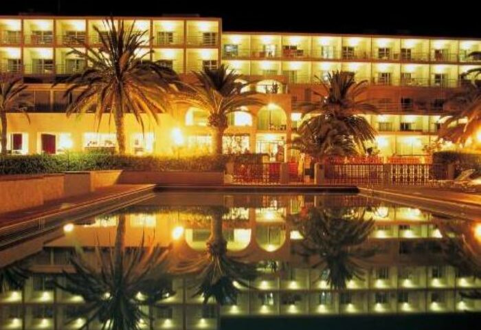 Hotel Nautico Ebeso, a gay-friendly hotel, at night. Palm trees and building lights reflect in the pool. A great place for gay travel.