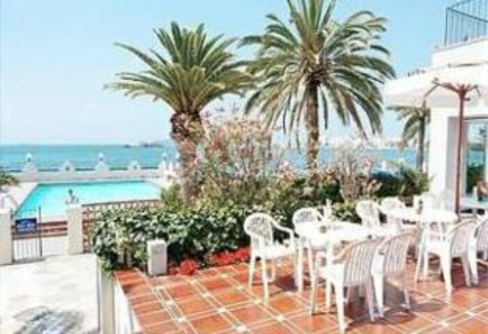 Hotel Nautico Ebeso, a gay-friendly hotel. Outdoor dining area with palm trees, pool, and ocean view. Perfect for gay travel in Ibiza.
