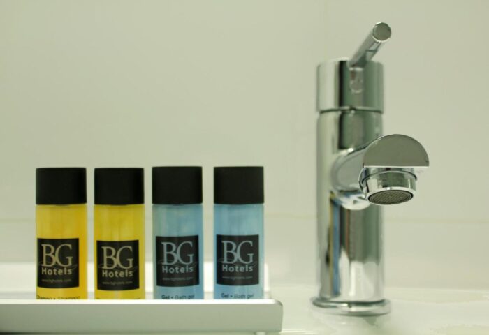 Hotel Nautico Ebeso's bathroom amenities. Yellow & blue BG Hotels shampoo & bath gel bottles next to a modern silver faucet. Gay-friendly hotel.