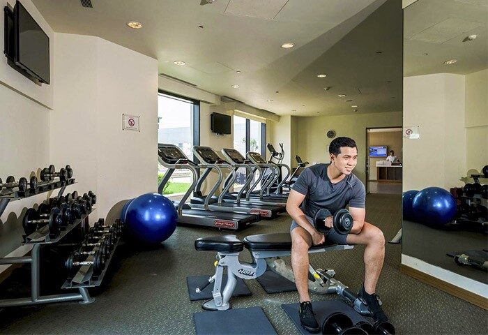 Hotel Nikko Saigon's gym: A gay-friendly hotel. Man lifting weights on a bench. Treadmills, dumbbells, and exercise ball are also visible.