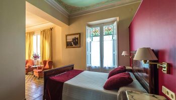 Hotel Noucentista's gay-friendly room: a bedroom with a double bed, a sitting area, and a window with colored glass. Gay travel.