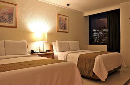Hotel PF's gay-friendly room featuring two beds, nightstand, lamp, and a window view of the city at night. Comfortable accommodations for gay travelers.