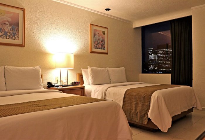Hotel PF's gay-friendly room featuring two beds, nightstand, lamp, and a window view of the city at night. Comfortable accommodations for gay travelers.
