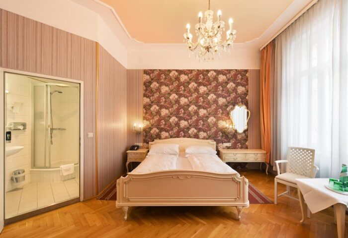 Hotel Pensione Wildner's gay-friendly room. A well-lit bedroom with a floral accent wall, chandelier, and a modern bathroom with a glass-enclosed shower.