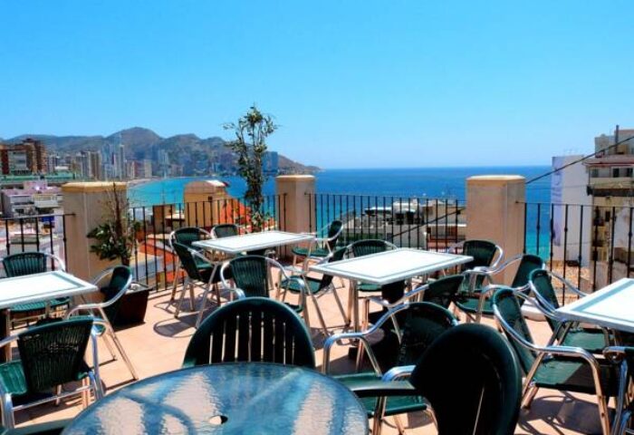 Rooftop terrace at Hotel Queens Adults Only By Mc, a gay-friendly hotel. Tables and chairs overlook the ocean and city skyline.