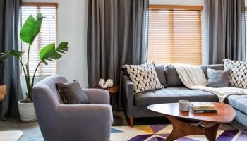 Hotel Royal gay-friendly hotel room: Gray couch, chair, and curtains. Geometric rug, coffee table, and potted plant. Window with blinds.
