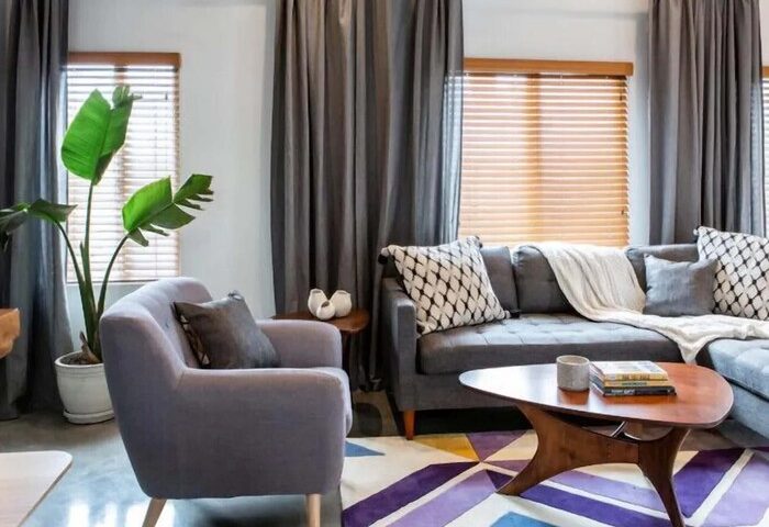 Hotel Royal gay-friendly hotel room: Gray couch, chair, and curtains. Geometric rug, coffee table, and potted plant. Window with blinds.