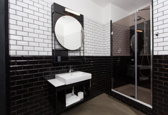 Hotel Rum Budapest gay-friendly bathroom. Black and white tiled walls, modern sink, round mirror, and glass-enclosed shower. Stylish design.