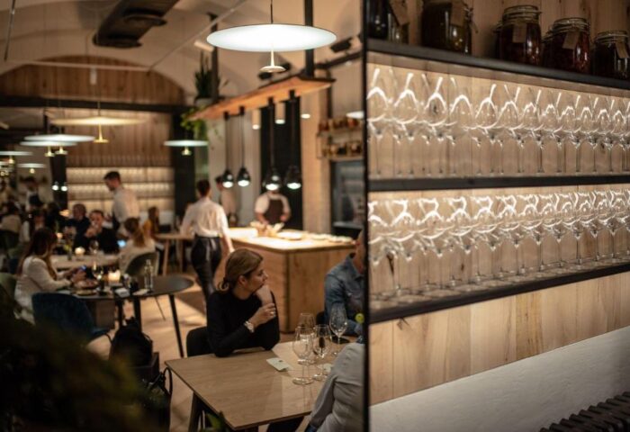 Hotel Rum Budapest's restaurant. Guests dine under modern lighting. Wine glasses line shelves. A gay-friendly hotel with a stylish bar.