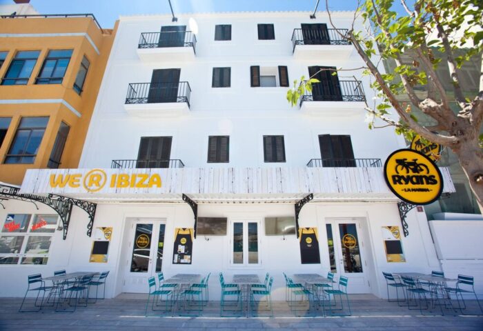 Ryans La Marina Hotel, a gay-friendly hotel in Ibiza. The hotel's exterior is white with black shutters and balconies. Tables outside.