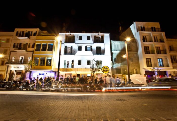 Hotel Ryans La Marina at night. Gay-friendly hotel with vibrant nightlife. Street view with people, motorbikes, and light trails in Ibiza.