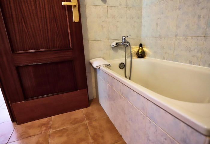 Bathtub in a room at Hotel Santa Faz, an adults-only, gay-friendly hotel. The tub has a shower head and tiled surround. Brown door and floor.