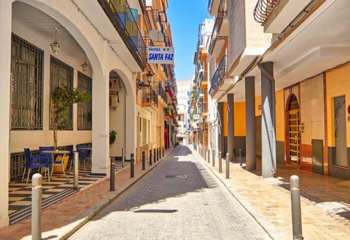 Street view of Hotel Santa Faz, an adults only, gay-friendly hotel. Cobblestone street, buildings on either side, blue sky.