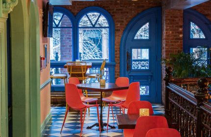 Hotel Scherman, a gay-friendly hotel. Interior view with red chairs, tables, and a black-and-white checkered floor. Blue door and windows.