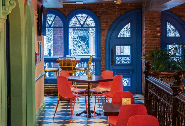 Hotel Scherman, a gay-friendly hotel. Interior view with red chairs, tables, and a black-and-white checkered floor. Blue door and windows.