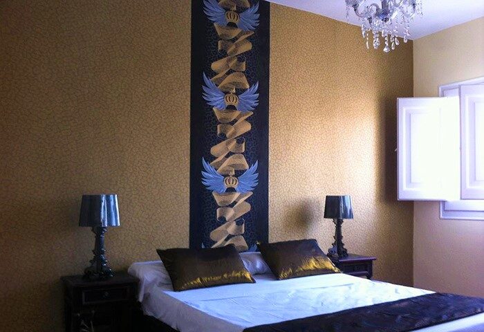 Hotel Sitges 1883 gay-friendly room. Bedroom with double bed, bedside tables with lamps, chandelier, and decorative wall art. Gay travel.