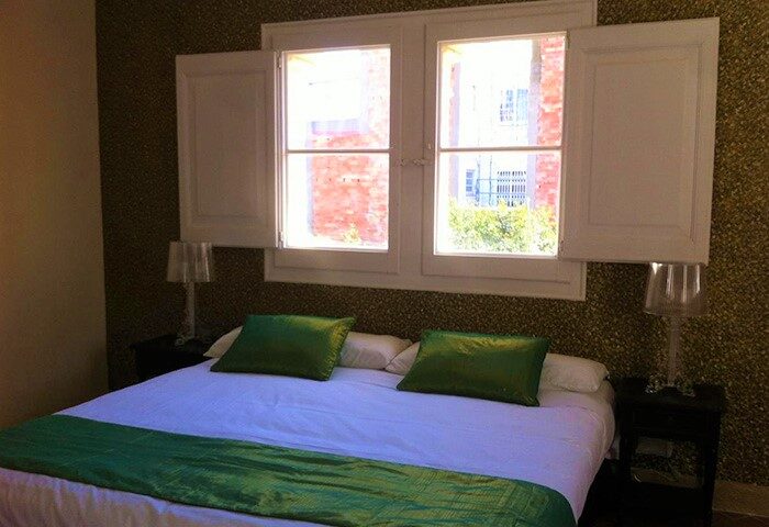 Hotel Sitges 1883 gay-friendly room. Double bed with green accents, bedside tables, lamps, and window with open shutters. Relaxing stay.