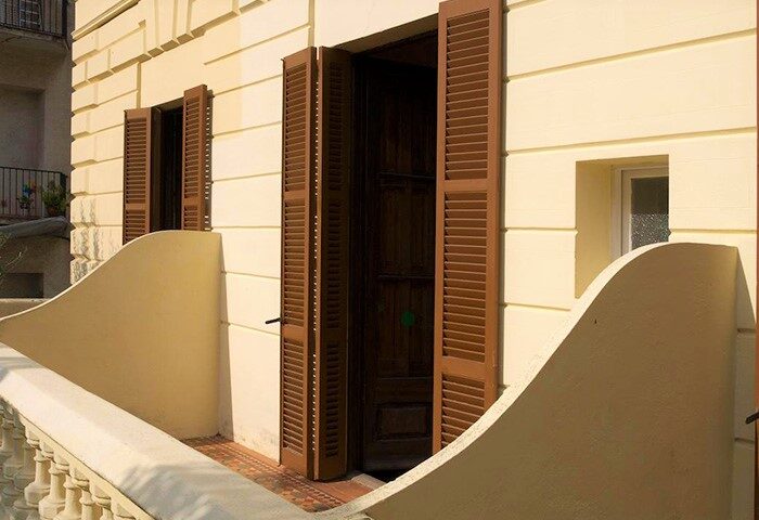 Exterior of Hotel Sitges 1883, a gay-friendly hotel. Balcony with brown shutters open to a dark doorway. Sunny yellow facade.